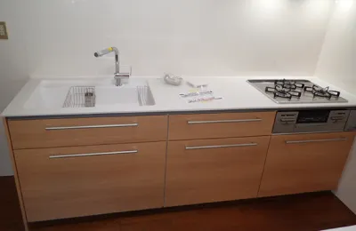 木目調の収納扉と白い天板を備えたシステムキッチンで、シンクとガスコンロ、ビルトイン機器が横並びに配置された様子