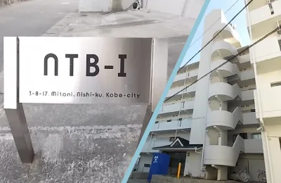 建物名「NTB-I」と所在地が記された金属製の銘板と、背後に立つ集合住宅の外観を組み合わせた写真