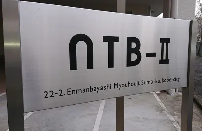 建物名「NTB-Ⅱ」と所在地が記された金属製の看板を正面から撮影した写真で、集合住宅の敷地入口に設置されている様子