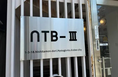 建物名「NTB-Ⅲ」と所在地が記された金属製の看板を正面から写した写真で、縦格子の門扉に取り付けられている様子