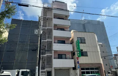 周囲に工事用の養生シートが張られた建物が並ぶ街並みで、中央に集合住宅が立つ外観写真